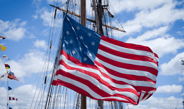 An American flag waves in the wind with a tall ship's masts and rigging in the background under a partly cloudy sky.