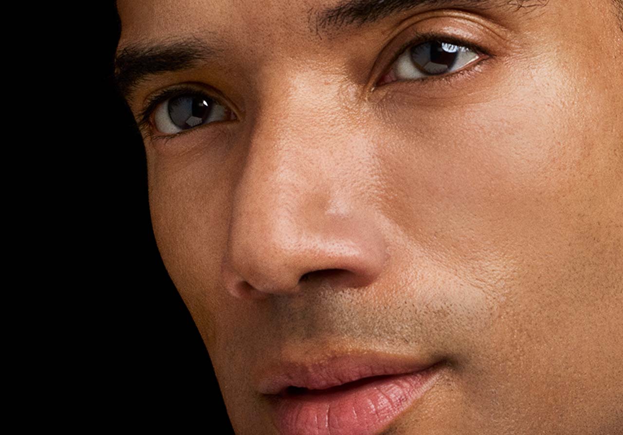 Nahaufnahme des Gesichts eines Mannes mit warmem, mittlerem Hautton vor schwarzem Hintergrund, mit Fokus auf Augen, Nase und Mundpartie.