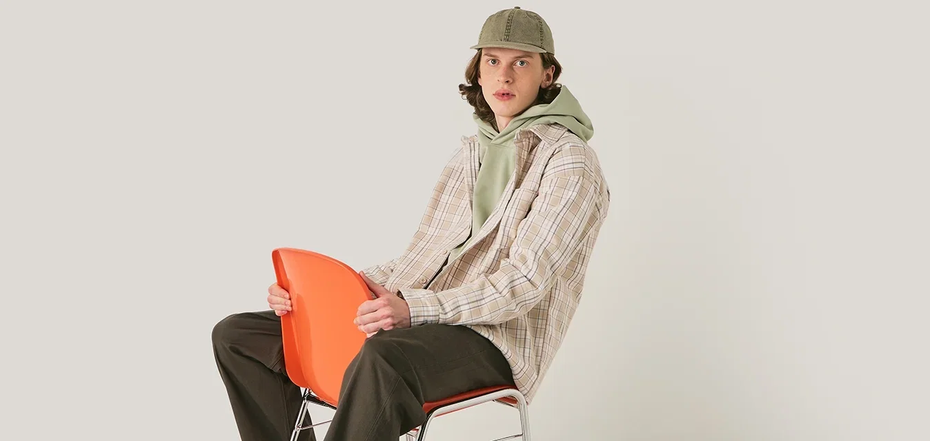 Geruite shirt in beige, groene hoodie, donkere broek, zwarte schoenen, houdt een oranje stoel vast. Glad textuur, modern ontwerp, neutrale achtergrond.