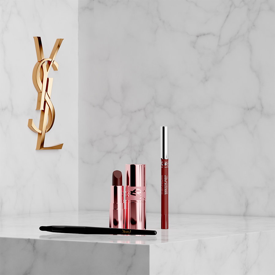 A YSL lipstick in a metallic rose gold case, a lip liner, and a black brush displayed on a white marble pedestal next to a golden YSL logo.