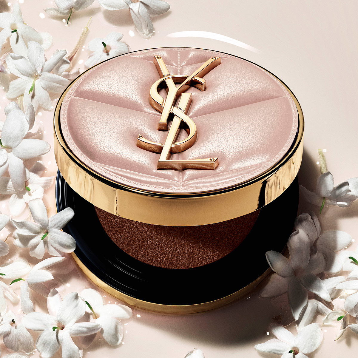 A YSL cushion foundation in a quilted pink and gold case, surrounded by delicate white jasmine flowers.