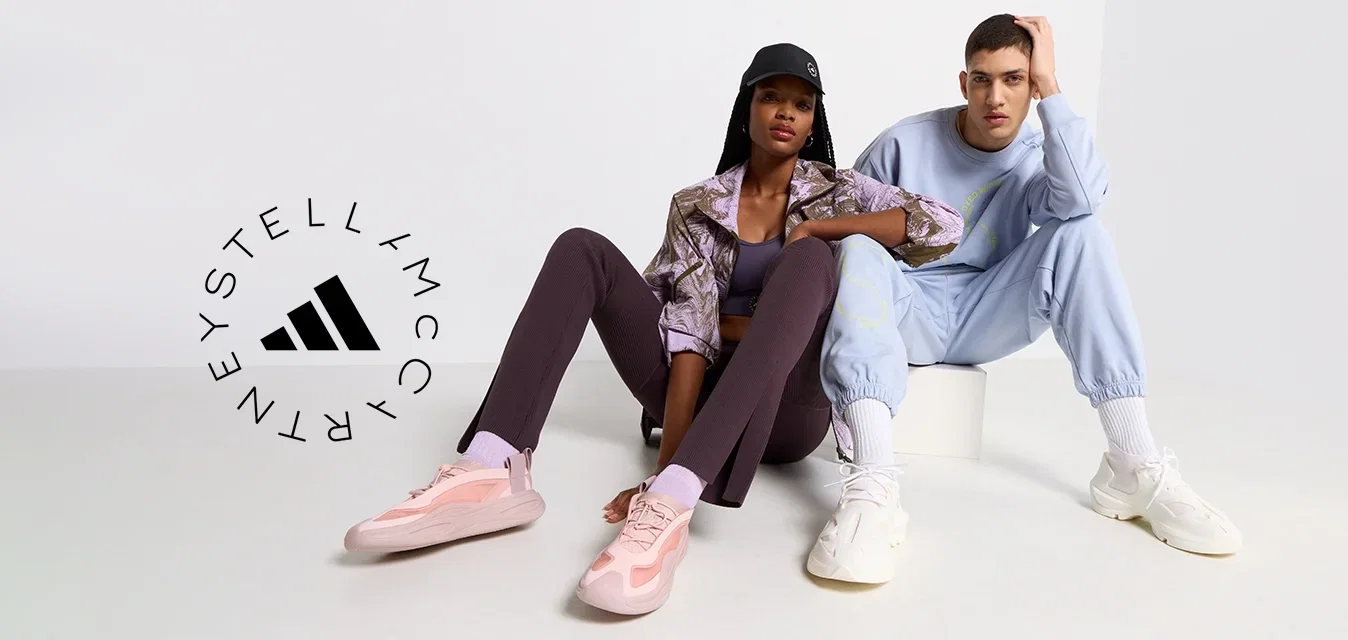 Zwei Models sitzen auf einer hellen Fläche. Eine trägt eine gemusterte braune Jacke, lila Leggings und rosa Sneaker. Die andere trägt einen hellblauen Trainingsanzug und weiße Sneaker.
