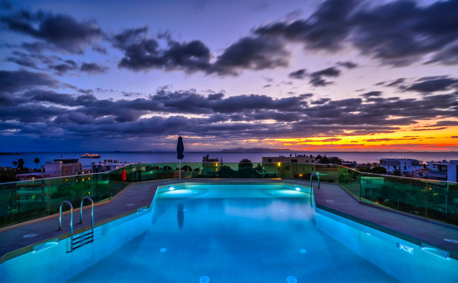 Rooftop infinity pool lit blue overlooking the sea and a cruise ship at colourful sunset with dramatic cloud-filled sky