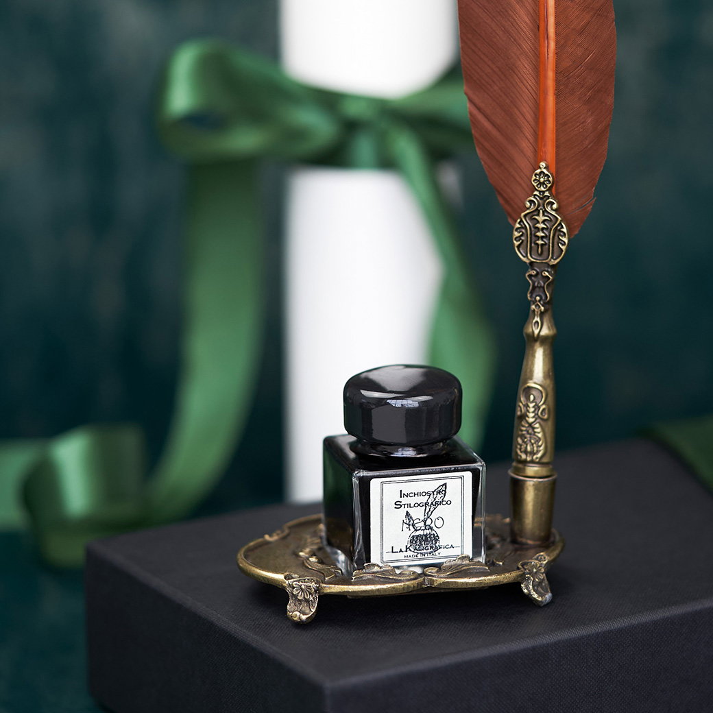 Calligraphy feather pen with ornate pewter handle and black ink bottle on a matching stand, arranged on a gift with tied ribbon in the background. - Graduation Gifts.