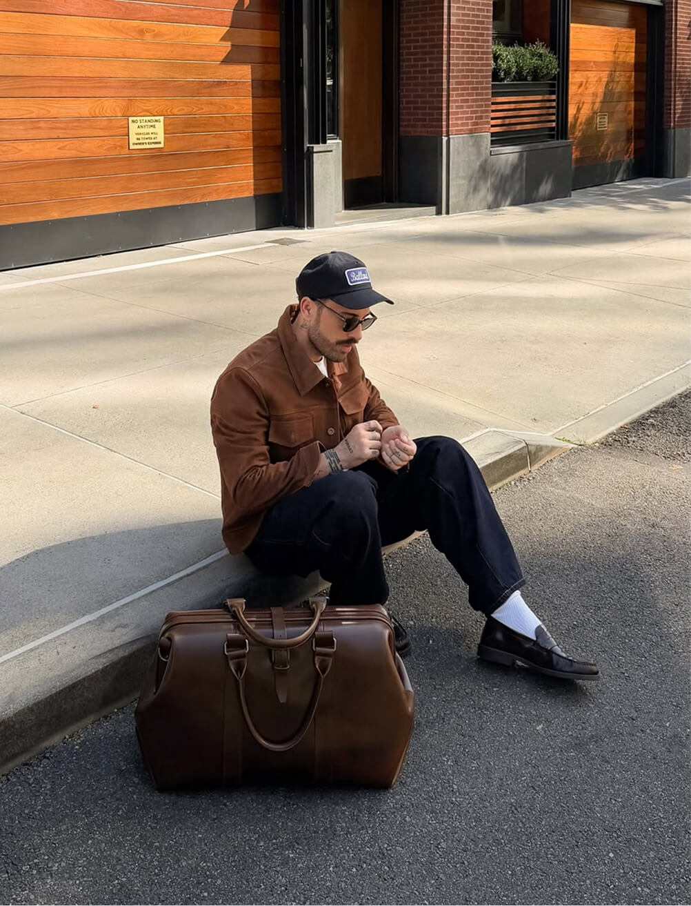 A man sits on a curb outside a building with wood-paneled and brick facade, looking down at his hands. He wears a black logo patch cap, narrow sunglasses, a brown suede overshirt, black trousers, white crew socks, and black loafers. A large structured brown leather duffle bag with top handles and buckle hardware sits on the street in front of him.