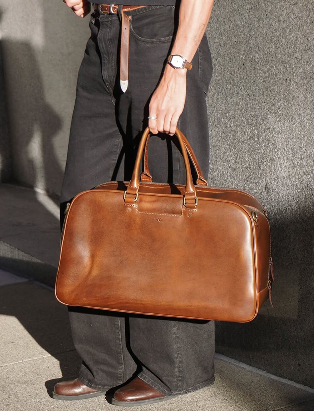 Close-up of a man holding a large structured tan leather duffle bag with dual top handles, brass hardware, and contrast stitching, photographed against a concrete wall. He wears dark charcoal jeans, brown leather boots, and a silver watch.