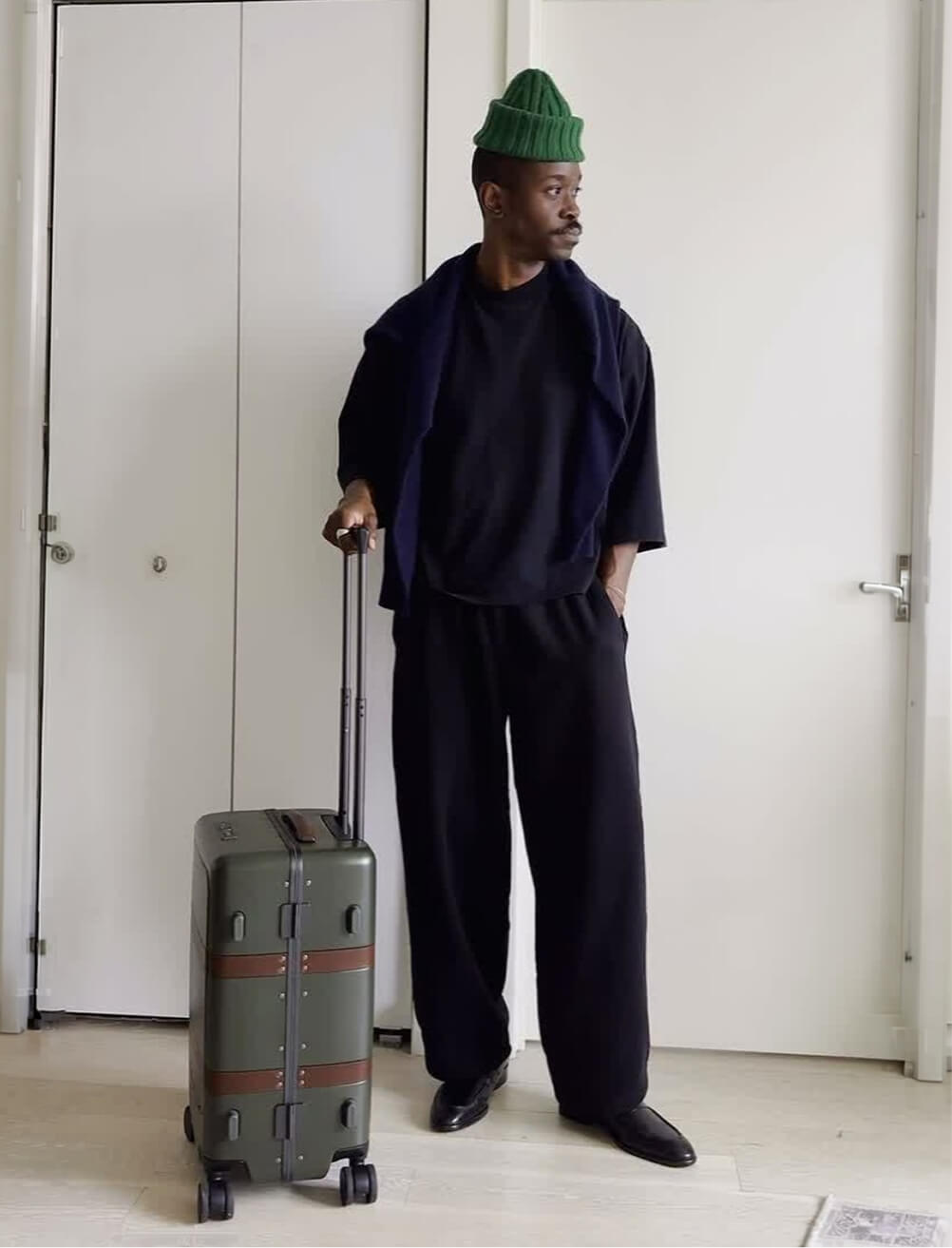 A man stands in a stylishly furnished interior space with a geometric mosaic tile floor, holding a green canvas and brown leather briefcase at his side. He wears a navy linen blazer over a white t-shirt and wide-leg olive trousers, with sunglasses pushed up on his head. He looks to the side, with one hand in his pocket.