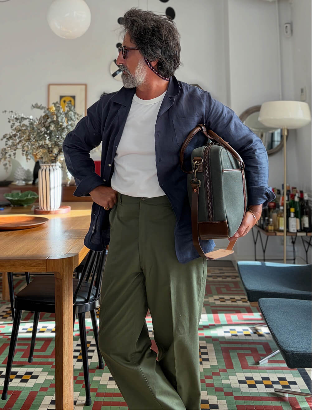 A man stands in a stylishly furnished interior space with a geometric mosaic tile floor, holding a green canvas and brown leather briefcase at his side. He wears a navy linen blazer over a white t-shirt and wide-leg olive trousers, with sunglasses pushed up on his head. He looks to the side, with one hand in his pocket.