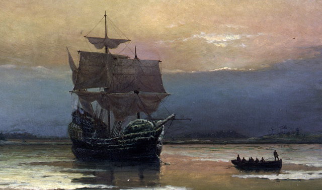 A historic sailing ship with large sails is anchored near the shore under a cloudy sky, with a small rowboat approaching it.