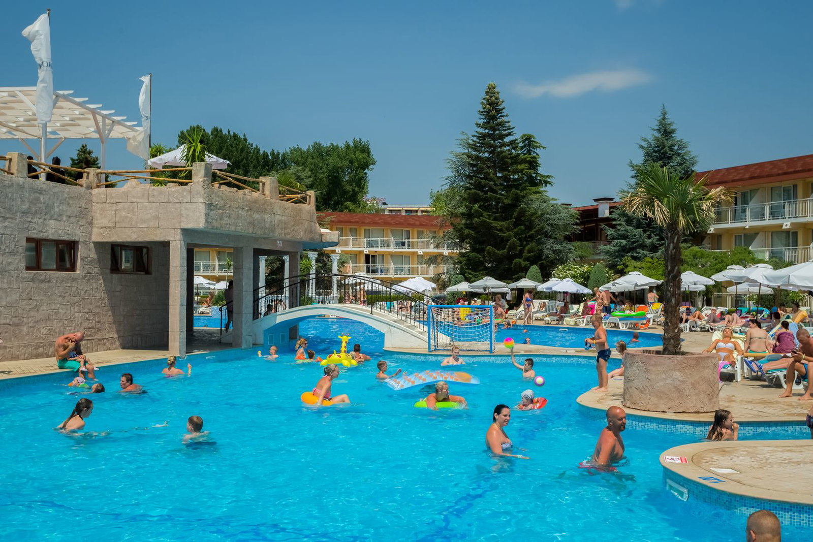 Outdoor resort hotel swimming pool with families and sunbathers, bridge, palm trees and umbrellas under a clear blue summer sky