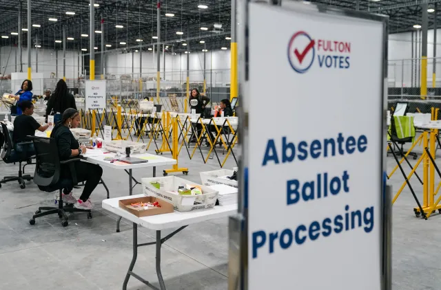 A large indoor space with workers at tables and temporary barriers, with a sign reading 