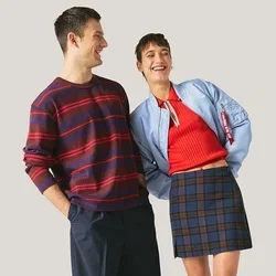 Man in een gestreepte trui en donkere broek staat glimlachend naast een vrouw in een rood topje, blauwe jas, geruite rok en zwarte laarzen tegen een neutrale achtergrond.