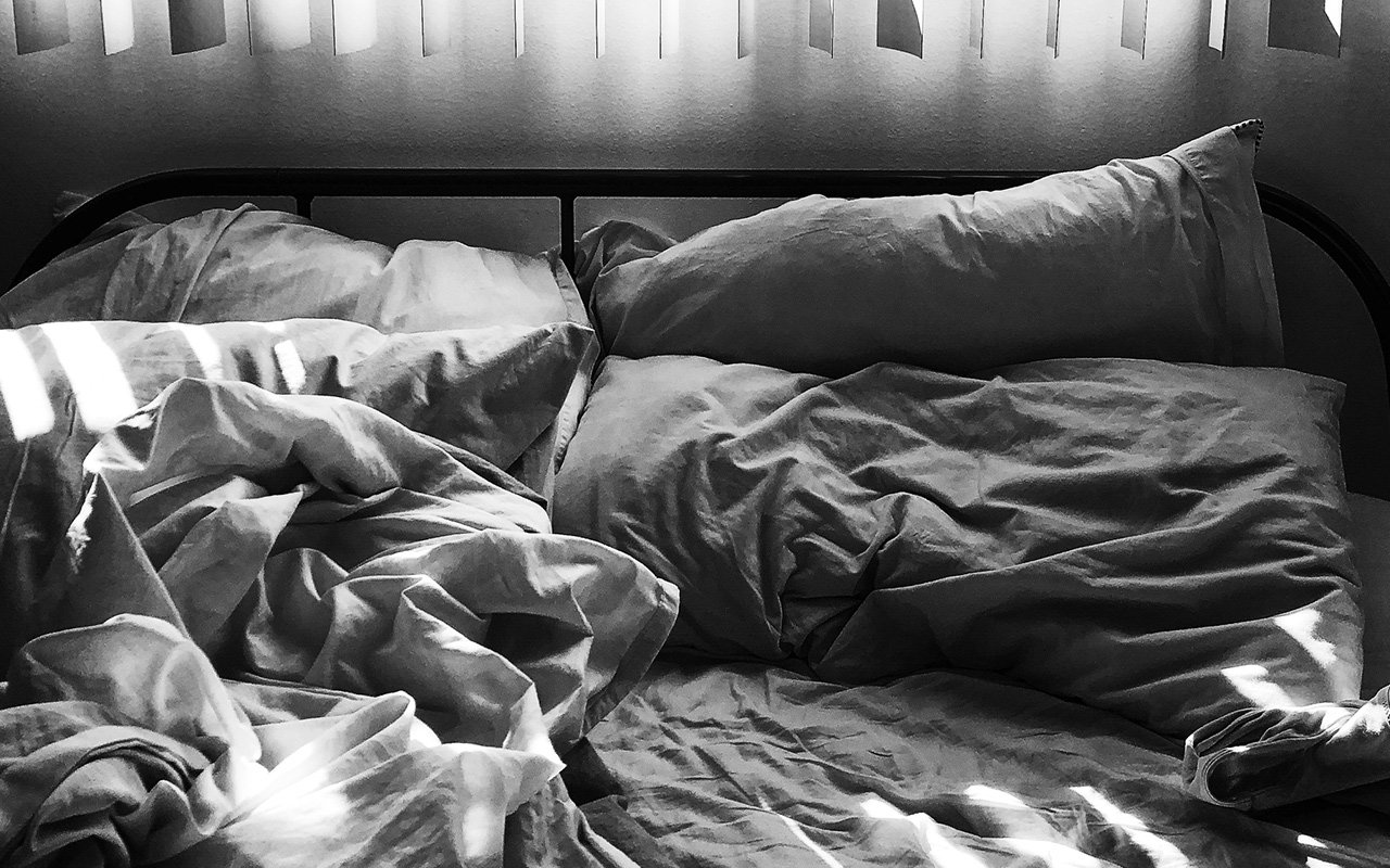 A rumpled bed with white sheets and pillows is lit by sunlight streaming through vertical blinds.