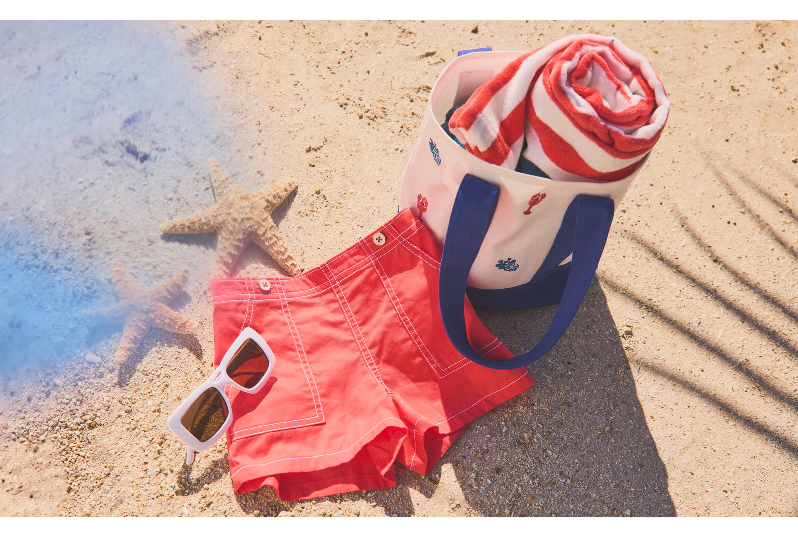 Beach scene with red swim shorts, white sunglasses, striped towel in beach bag, and starfish on sand
