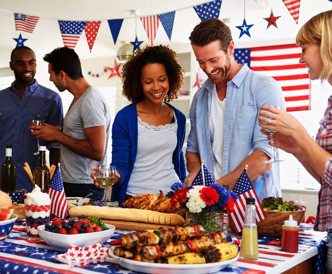 Friends gather around a Fourth of July table with grilled food, drinks and patriotic decorations