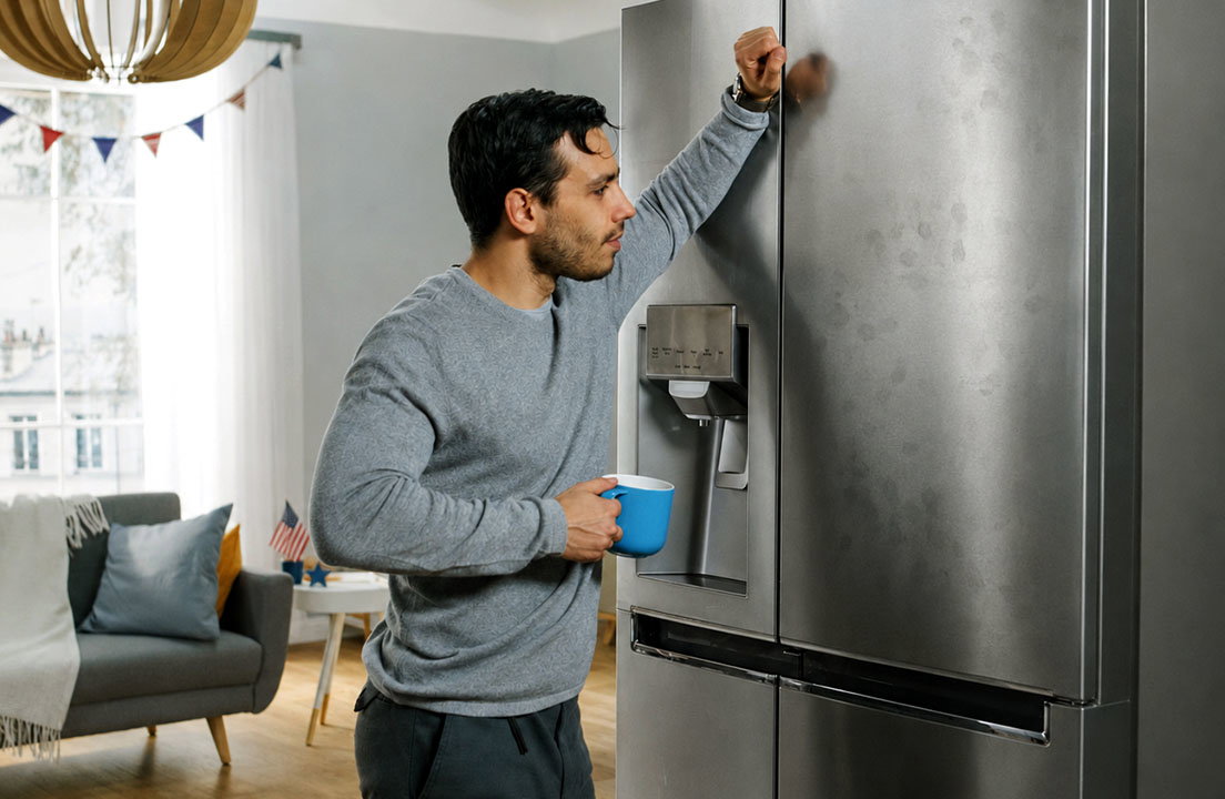 A man fills a mug from a stainless steel refrigerator with visible fingerprints on the door