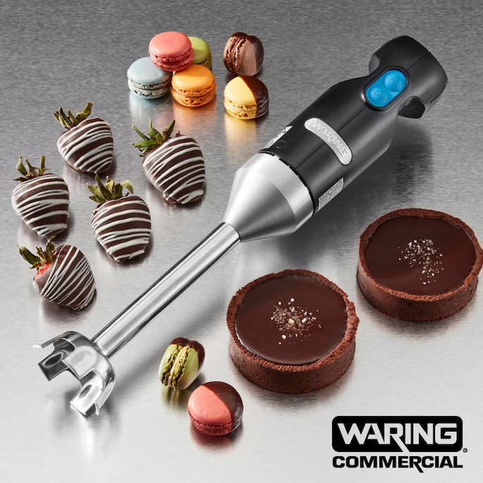 Waring Commercial immersion blender displayed with chocolate-covered strawberries, macarons, and chocolate tarts on a stainless steel surface.