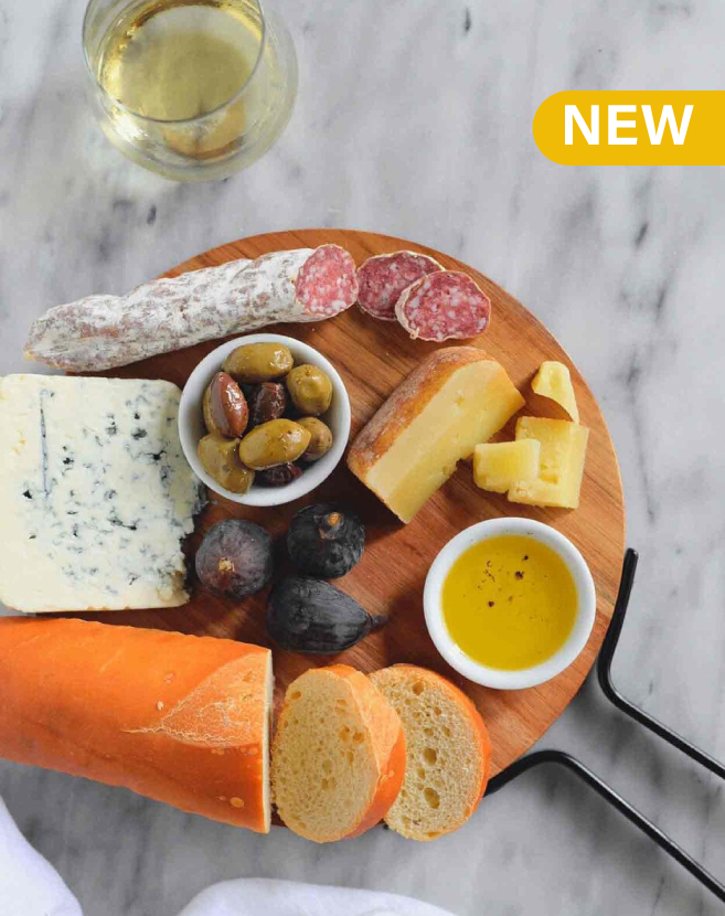 Round wooden serving board featuring assorted cheeses, cured meats, olives, bread, figs, and olive oil on a marble surface, with a New Items banner displayed above.”