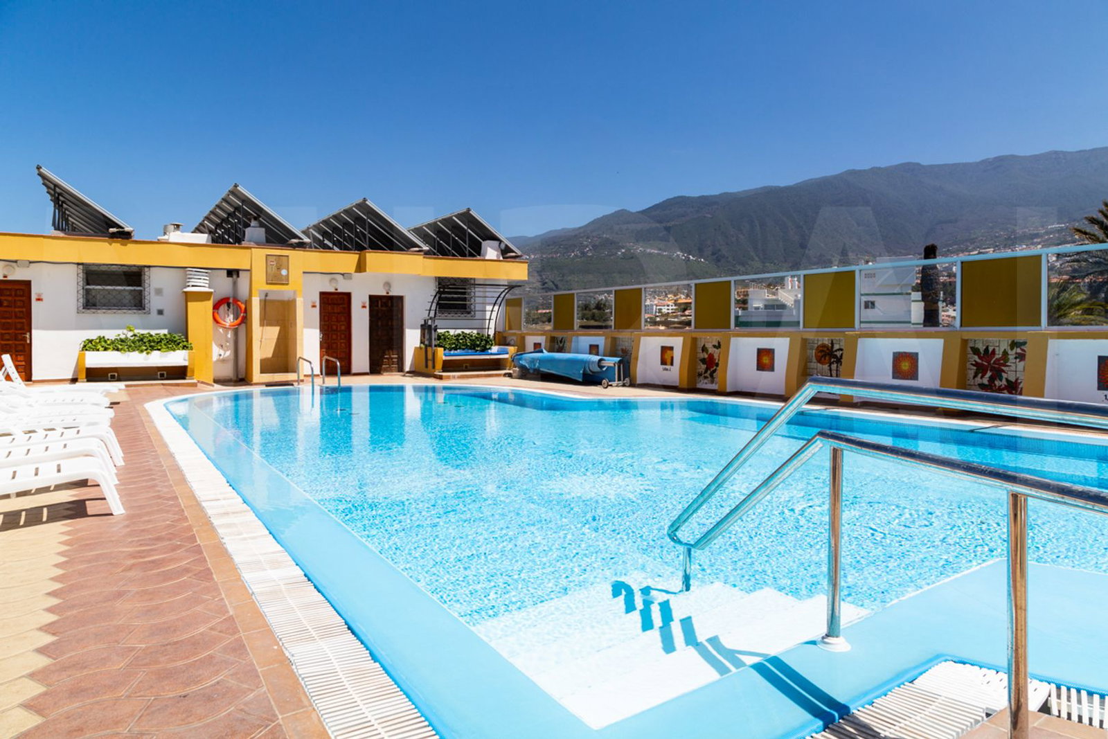 Rooftop hotel swimming pool with clear blue water, sun loungers, stainless steel handrail and mountain view under a bright blue sky