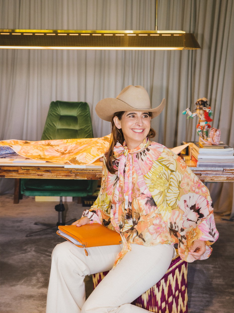 Designerin Dorothee Schumacher sitzt lächelnd in ihrem kreativen Atelier. Sie trägt eine farbenfrohe Seidenbluse mit floralem Print, weiße Hosen, einen beigen Cowboyhut und hält ein orangefarbenes Leder-Portfolio.