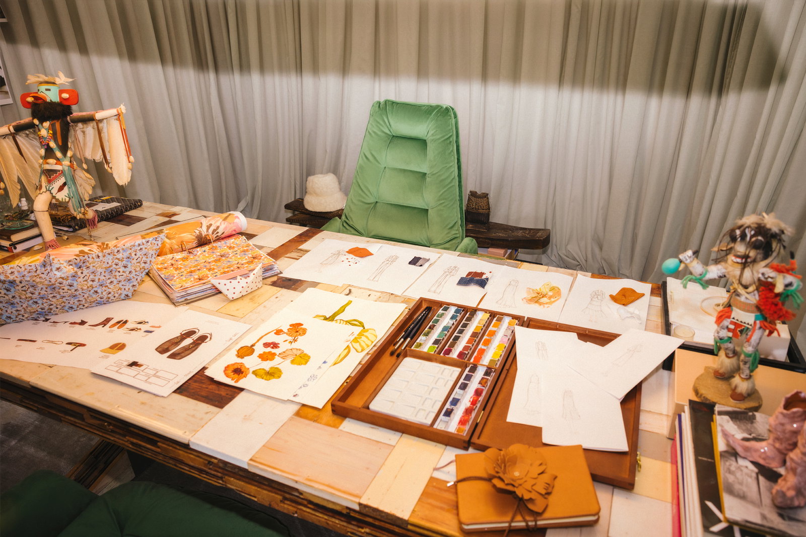 Ein Blick auf den inspirierenden Designtisch im DOROTHEE SCHUMACHER Atelier, bedeckt mit Skizzen, Stoffproben, einem Aquarell-Malkasten und kunstvollen Skulpturen vor einem grünen Samtsessel.