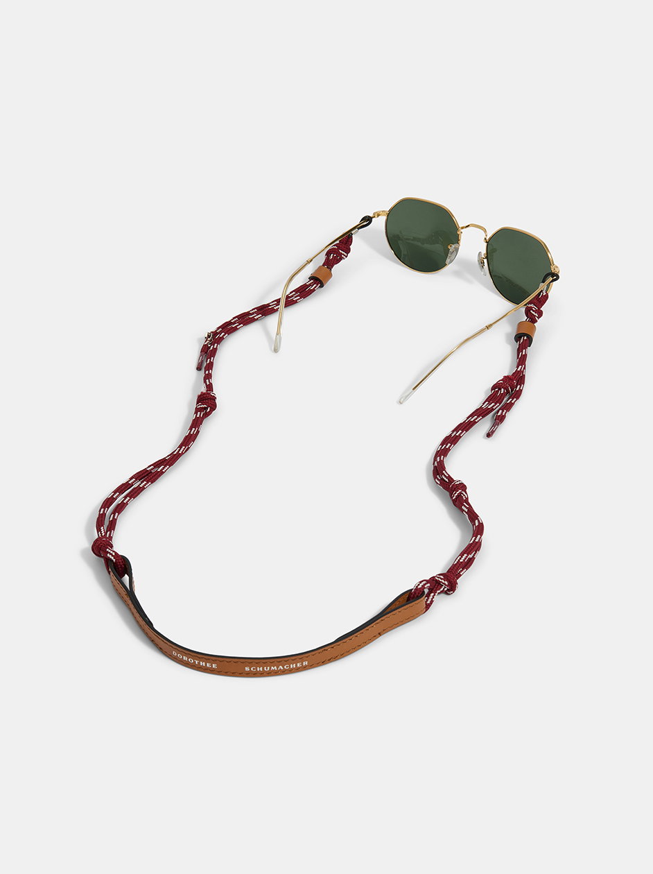 Eine runde Sonnenbrille mit goldenem Gestell von DOROTHEE SCHUMACHER, befestigt an einer sportlichen, rot-weiß gemusterten Kordel-Brillenkette mit einem geprägten braunen Lederband im Nacken.