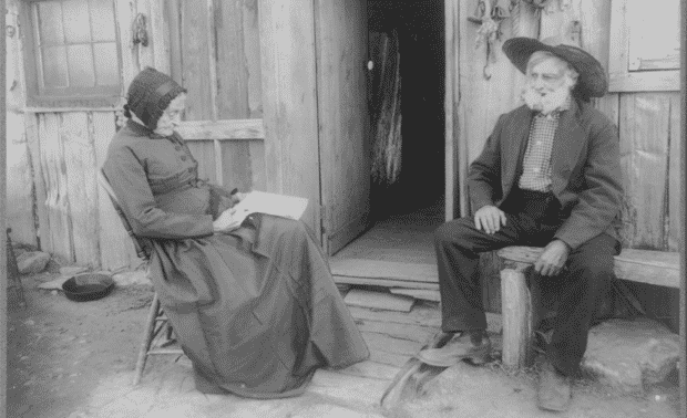An elderly woman and man sit outside a rustic wooden building. The woman is reading a book while the man, wearing a large hat, looks on.