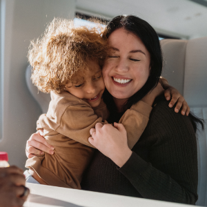 A parent sits by a window on a train while holding a young child in a warm embrace.