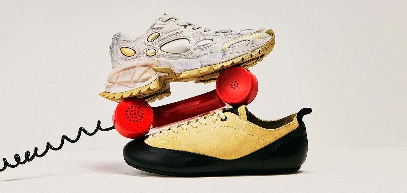 Rode vintage telefoonhoorn gebalanceerd tussen twee sneakers, de ene beige en zwart en de andere lichtgrijs met gele accenten, op een effen achtergrond.