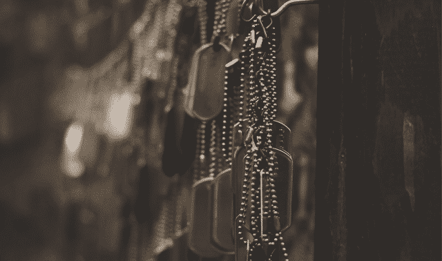 Close-up of hanging military dog tags on chains, displayed against a dark background. The tags are in focus with a blurred background effect.