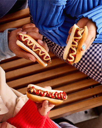 Three friends sitting on a bench holding hot dogs.