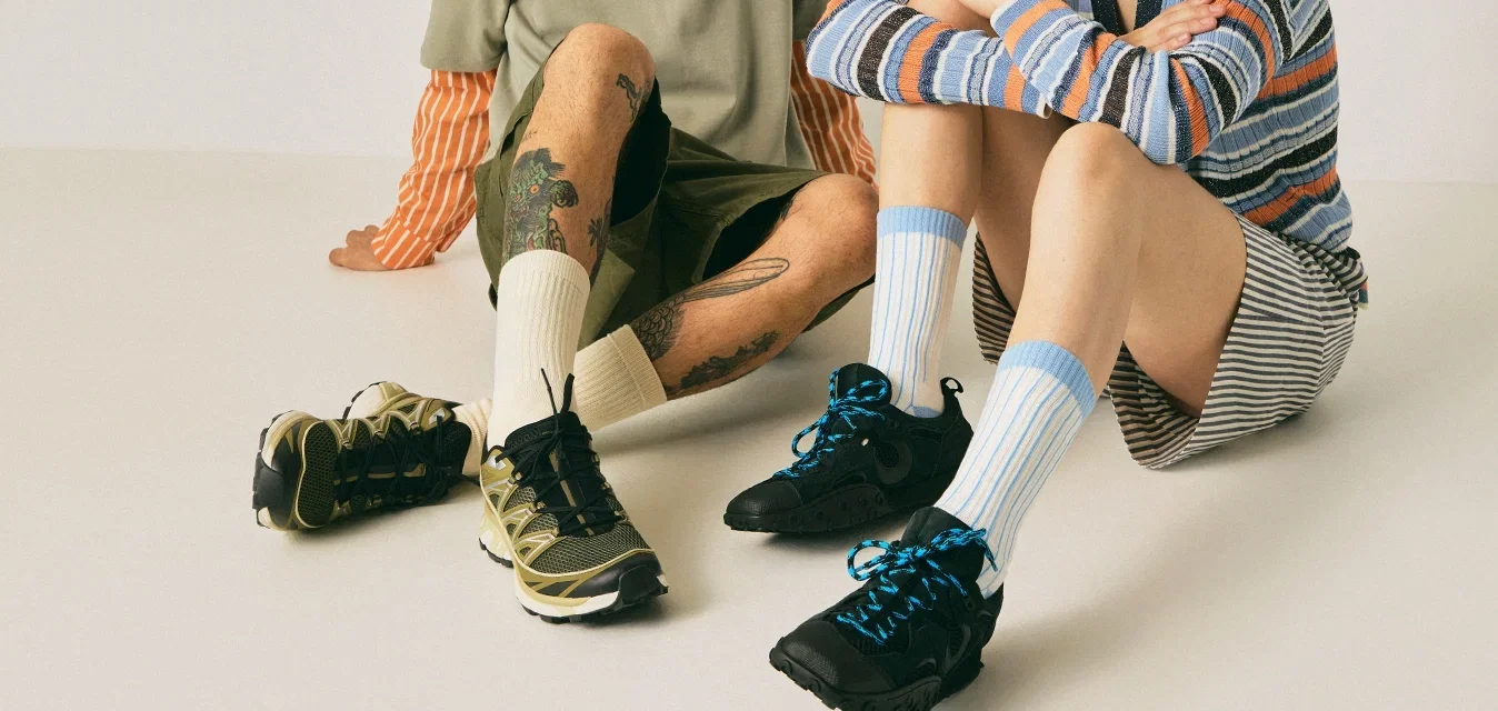 Twee mensen zitten op de vloer tegen een effen achtergrond, gekleed in casual gestreepte en effen kleding met sneakers en sokken.