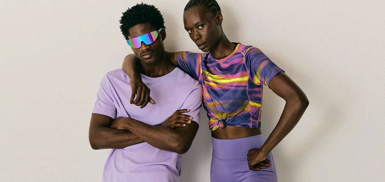 Twee volwassenen in paarse sportoutfits staan tegen een effen muur; de ene leunt met de armen gekruist, de andere heeft een hand op de heup en de arm rust op zijn partner.