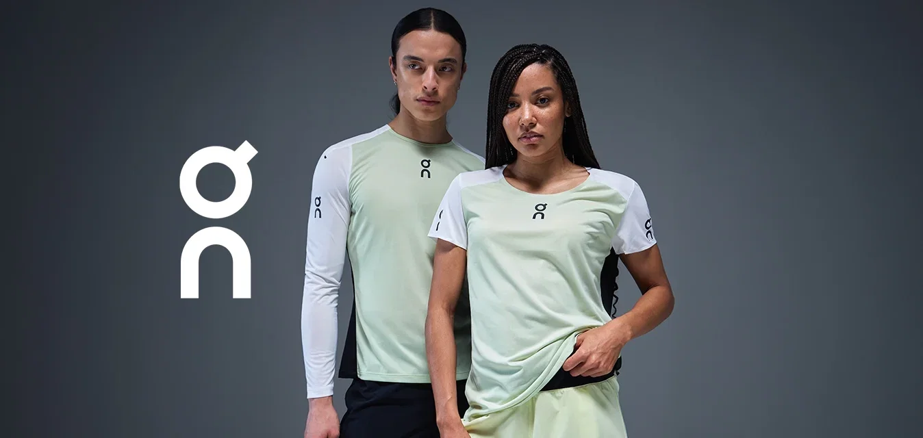 Twee modellen in lichtgroene en witte sporttops met logo's, die zelfverzekerd tegen een donkergrijze achtergrond staan.