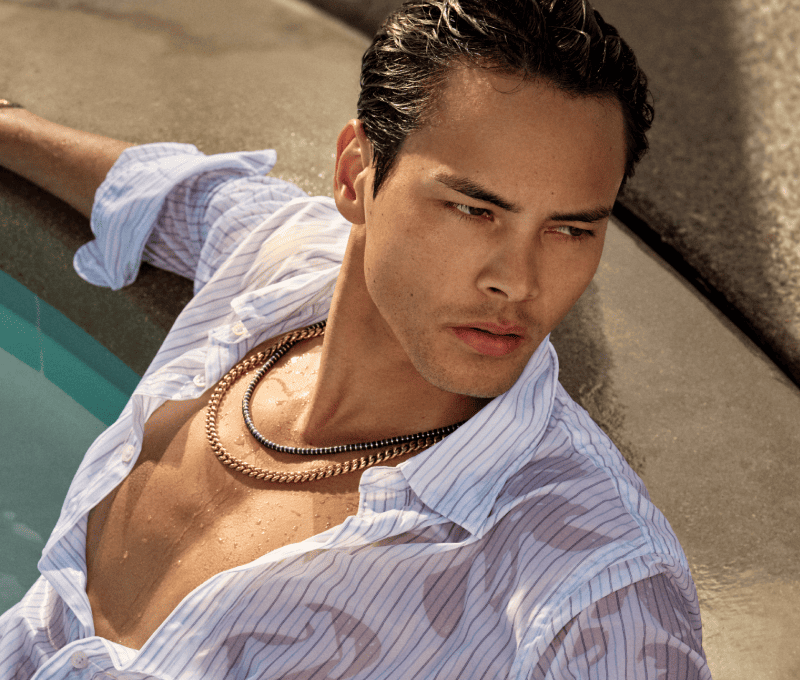 Male model reclining in pool wearing layered necklaces.