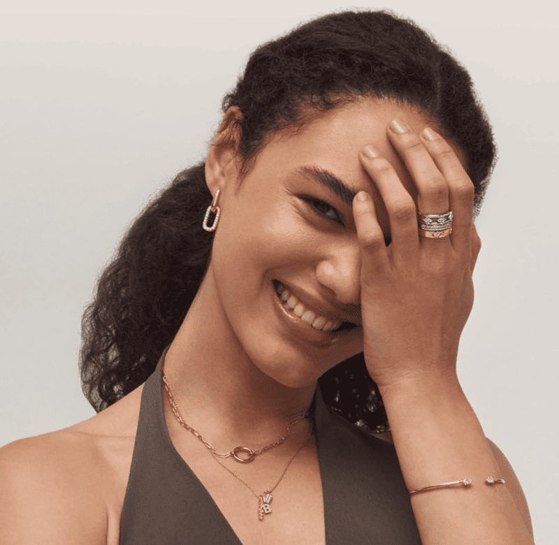 Woman wearing gold square hoop earrings, layered necklaces, a bangle, and a stack of rings. 