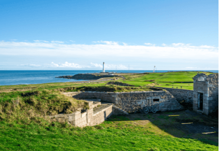Coastal landscape featuring grassy terrain, ruins, and a distant lighthouse under a clear blue sky.