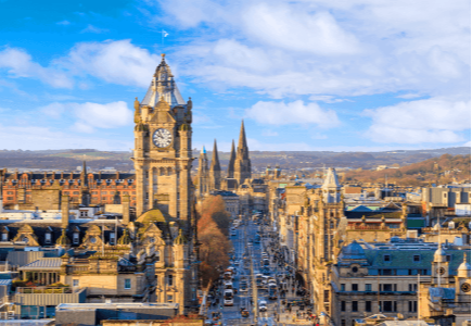 View of a cityscape featuring a clock tower, spires, and a bustling street under a blue sky.