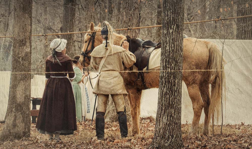 Two individuals in historical clothing stand beside a saddled horse in a wooded area, with ropes and a white tent in the background.