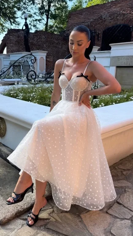 Woman in a white polka-dot tulle midi dress with corset bodice and black bows, seated on a stone wall in a garden courtyard