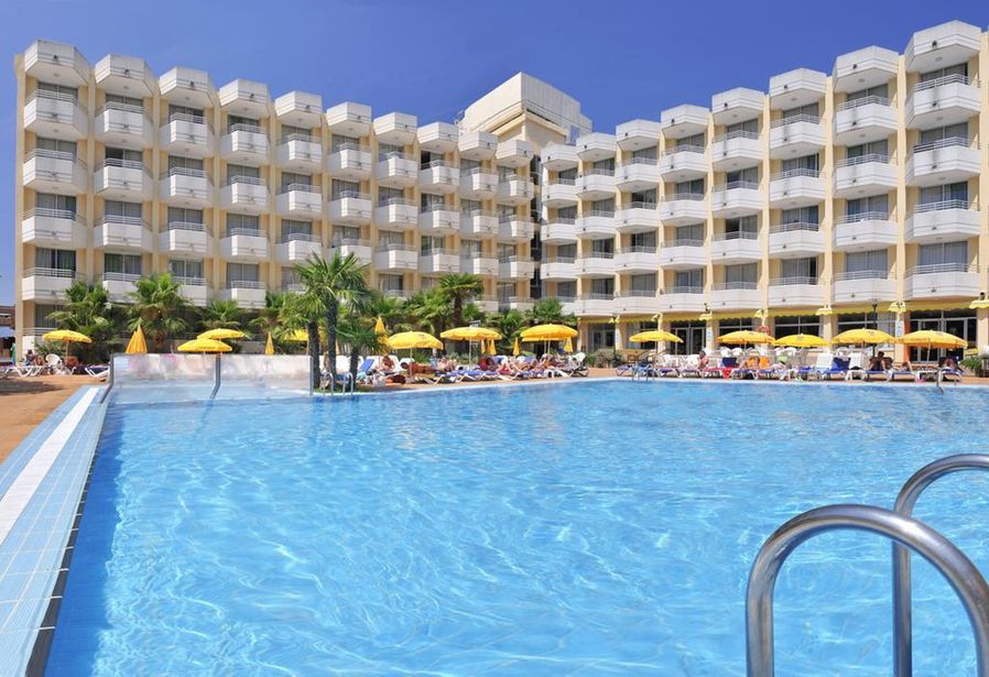 Large multi‑storey hotel with balconies beside a clear outdoor swimming pool, yellow parasols and sun loungers under a blue sky