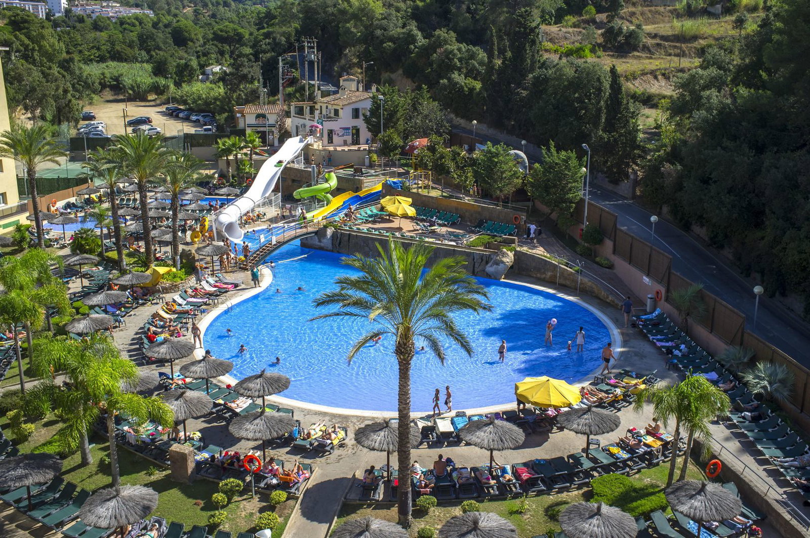 Aerial view of a resort swimming pool with water slides, palm trees, sun loungers and families enjoying a sunny holiday