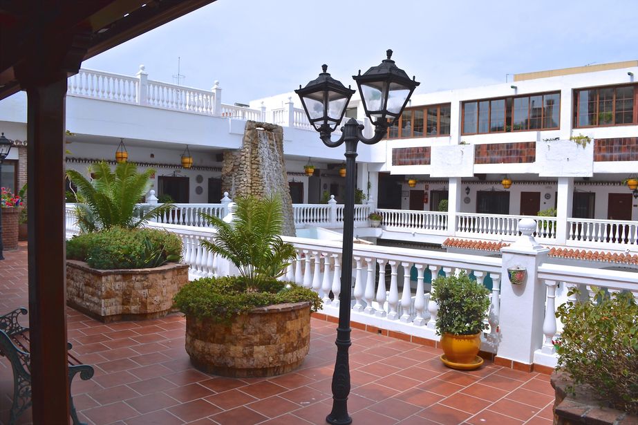 Hotel courtyard terrace with white balustrades, central black lamppost, terracotta tiles, potted palms and pool beyond