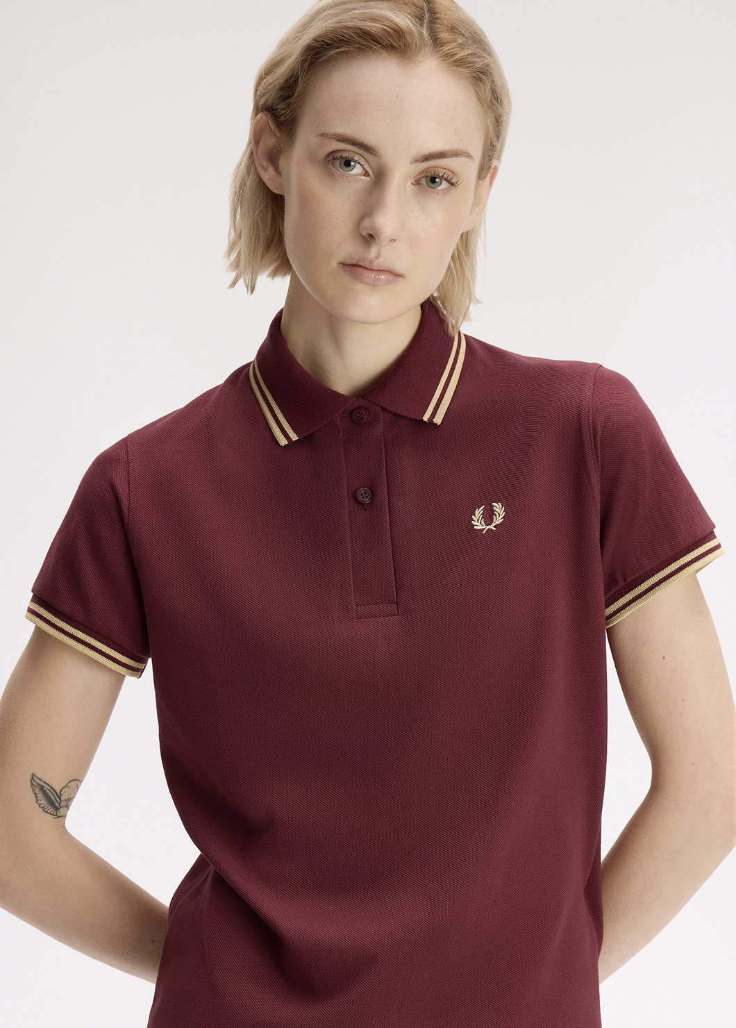 White female model with jaw-length blonde hair crosses her arms behind her back, stares straight into the camera and wears a Fred Perry G3600 Twin Tipped Shirt in Dark Red 