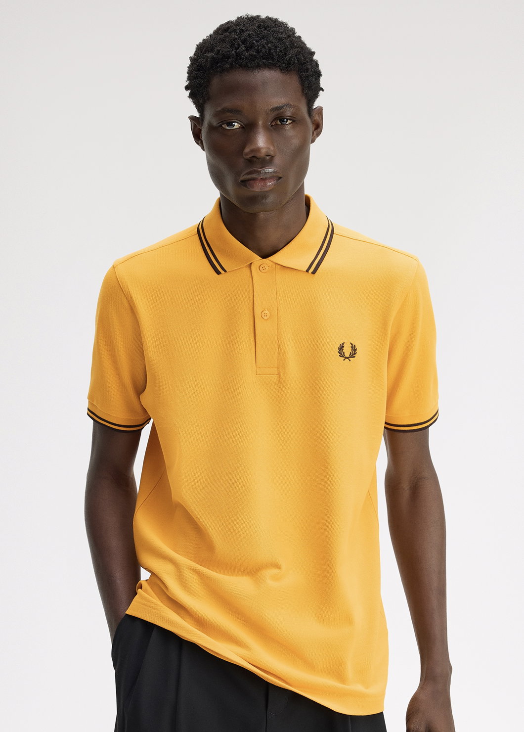 Black male model with short coily hair looks straight into the camera and wears a Fred Perry M3600 Twin Tipped Shirt in Golden / Rich Brown / Rich Brown, while he tucks one hand into his pocket