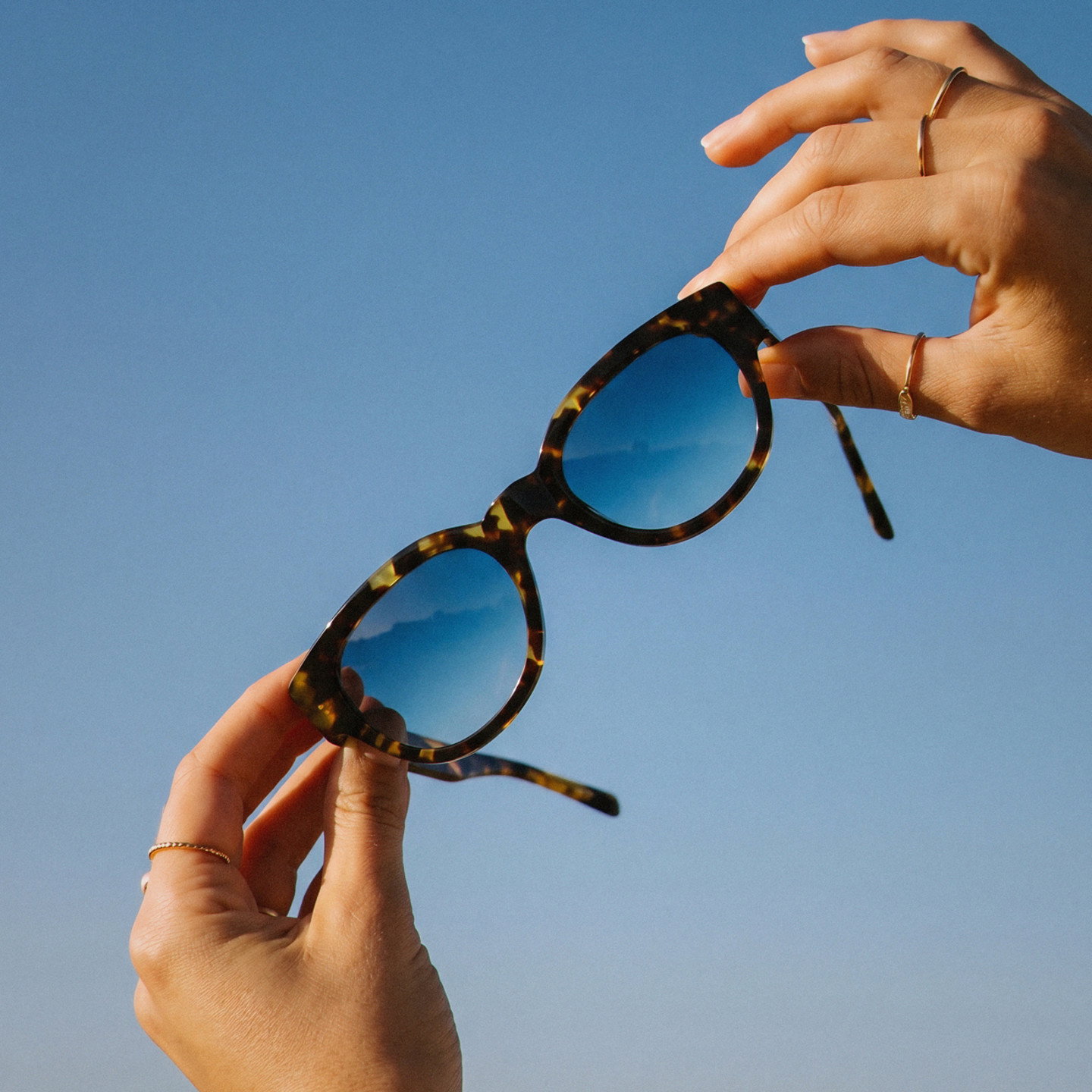 Eine Sonnenbrille mit Schildpattrahmen und blauen Verlaufsgläsern wird vor klarem Sommerhimmel gehalten.
