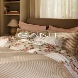 Slaapkamer met zonlicht, een netjes opgemaakt bed met gestreept en bloemig beddengoed, doorschijnende gordijnen, een bedlampje en een klein krukje met opgevouwen dekens.