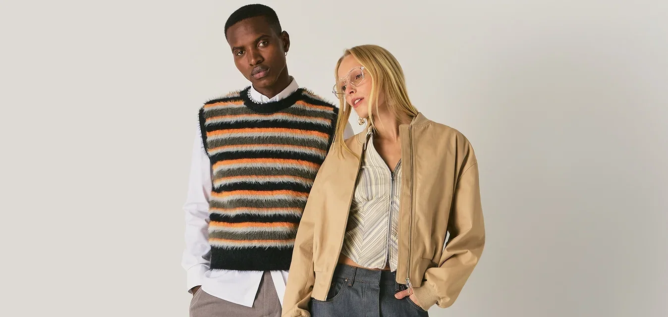 Twee modellen staan tegen een effen achtergrond, de een draagt een gestreept gebreid vest en wijde broek, de ander een beige jas, een patroonrok en laarzen.