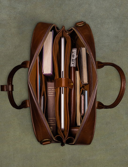 Overhead flat-lay of an open tan leather briefcase fully packed, showing a laptop secured in a center strap, two books, a USB hub, a smartphone, pens, and a small notebook organized across multiple interior compartments and pockets.