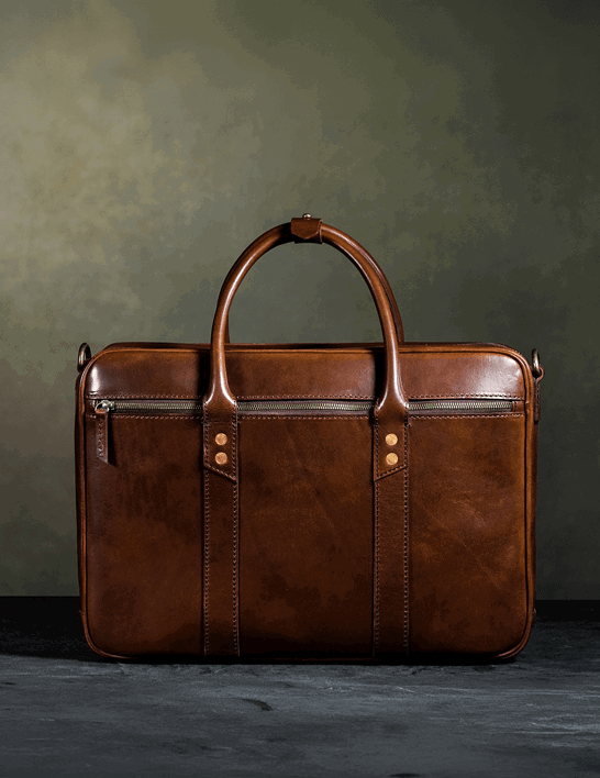 Front view of a structured tan leather briefcase with dual top handles, contrast stitching, a front zip pocket, vertical panel detailing, and copper rivet accents, photographed against a muted olive green background.
